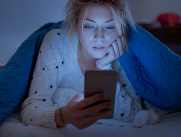 Frau mit blonden Haaren sitzt im Dunklen auf ihrem Bett und wird nur von dem blauen Licht ihres Smartphones angestrahlt