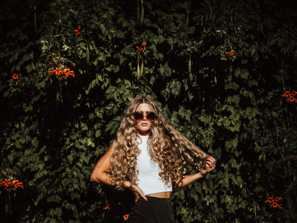 junge Frau mit langen, lockigen Haaren und einer Sonnenbrille steht vor einer Pflanzenwand