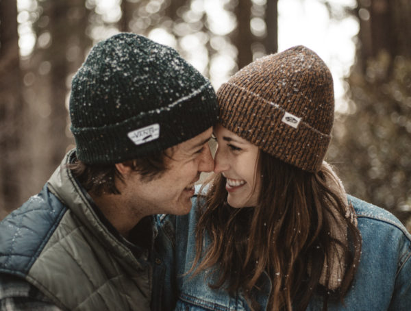 Nahaufnahme eines verliebten Paares im Wald. Beide tragen eine Mütze und blicken sich lächelnd an.