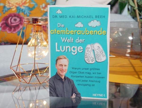 Buchcover von "Die atemberaubende Welt der Lunge" von Dr. med. Kai-Michael Beeh. Der Buchumschlag ist Türkis und der Autor ist auf dem Cover mit einer gezeichneten Lunge abgebildet. Das Buch steht auf einem Glastisch mit Deko-Elementen im HIntergrund.
