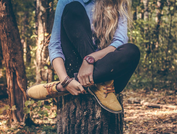 Frau mit schwarzer Jeans, blauem Jeanshemd und braunen Schuhen, die auf einem Baumstamm sitzt und die Beine vor dem Körper verschränkt. Das Gesicht ist nicht zu sehen aber lange blonde Haare, die sie über die linke Schulter trägt.