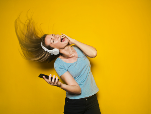 Frau hört über Kopfhörer Musik vor einem gelben Hintergrund und wirft dabei lachend ihre Haare zurück