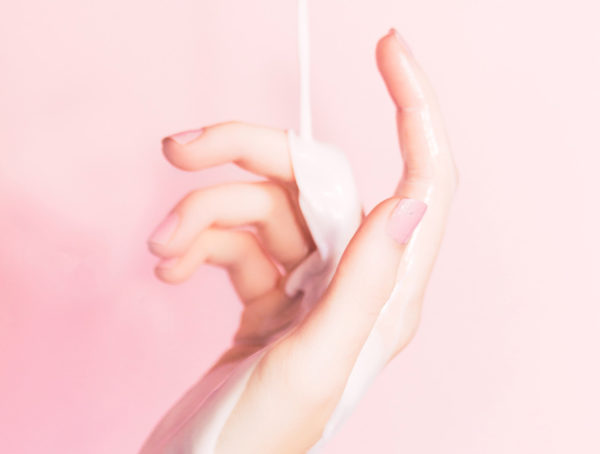 Hand mit rosa lackierten Nägeln vor rosa Hintergrund. Über die Finger läuft eine weiße Creme
