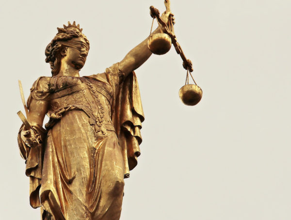 goldene Justitia mit verbundenen Augen und Waage vor einem hellen Hintergrund
