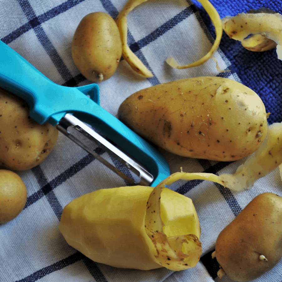 Kartoffeln werden mit einem Kartoffelschäler geschält und liegen auf einem blau-weißen Küchenhandtuch