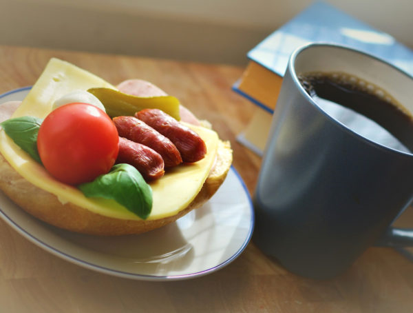 Brötchen mit Käse, Salmi, Tomate und Basilikum auf einem weißen Teller mit blauem Rand. Rechts daneben steht eine dunkelgraue Tasse mit schwarzem Kaffee, dahiner liegen zwei Bücher, die übereinander liegen