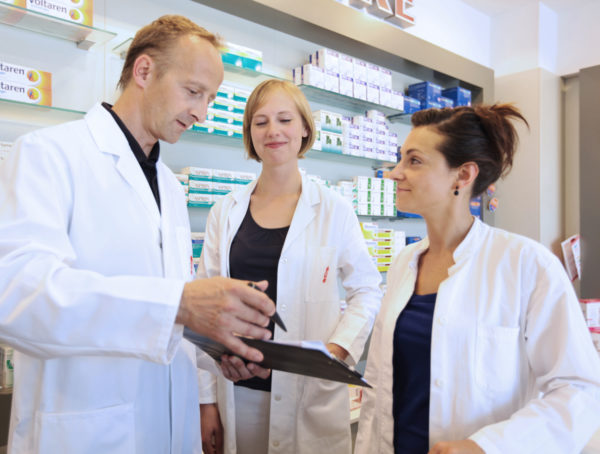 Apotheker und zwei PTA in weißem Kittel. Der Apotheker hält einen Stift und zeigt damit auf ein Papier Stimmungsbarometer