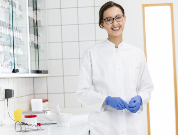 PTA mit Brille, braunen, Haaren, Kittel und blauen Handschuhen in der Rezeptur. Neben ihr stehen Fantaschale, Pistill und Waage.