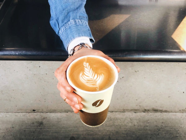 Kaffee in einem Kaffeebecher mit Bohne und braunem unteren Rand, der in einer Hand gehalten und über eine Tresen gereicht wird.