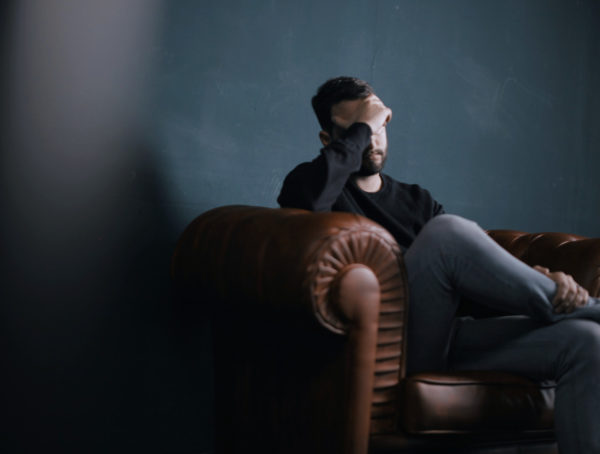 Mann mit Kopfschmerzen, der sich den kopf hält und auf einem braunen Ledersofa sitzt. Die Beine hat er überschlagen. Die Wand im Hintergrund ist dunkelblau