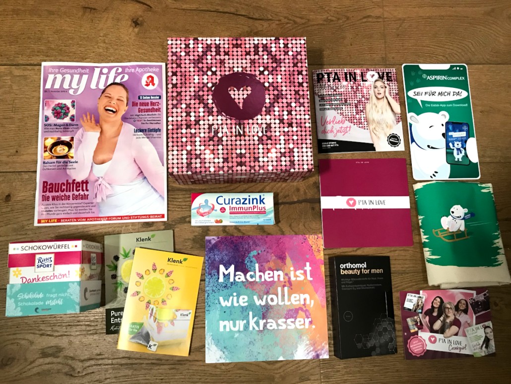 PTA IN LOVE Novemberbox mit ihrem Inhalt bestehend aus einem mylife Magazin, einem Rittersport Schokowürfel, zwei Teeproben von Klenk, einer Packung Curazink Immunplus, einer quadratischen bunten Karte mit der Aufschrift „Machen ist wie wollen, nur krasser“, dem PTA IN LOVE Magazin und den Produktkarten, einer Probe Orthomol Beauty for men, einem Turnbeutel mit dem Aspirineisbären sowie einer Karte in Smartphoneform mit dem Eisbären und einer Postkarte mit den PTA IN LOVE Covergirls