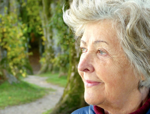 Ältere Dame mit grauen´m Haar, rotem Rollkragen und blauer Jacke, die von links nach rechts schaut. Hinter ihr ist ein Waldweg zu sehen.
