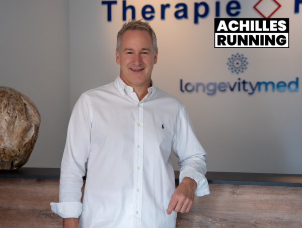 Ein Therapeut steht im Raum. Der Therapeut trägt ein weißes Hemd und lehnt sich am Counter. Was es damit auf sich hat, erfahrt ihr im neuen ACHILLES RUNNING Podcast.