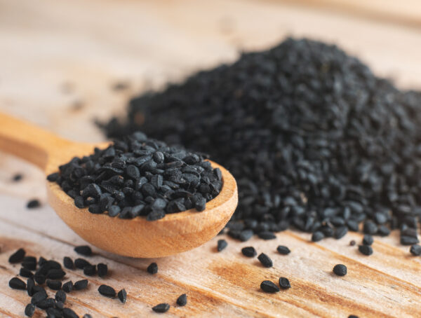Ein einem Holzlöffel liegen zahlreiche Schwarzkümmel-Samen, im Hintergrund ein Haufen mit Schwarzkümmel-Samen