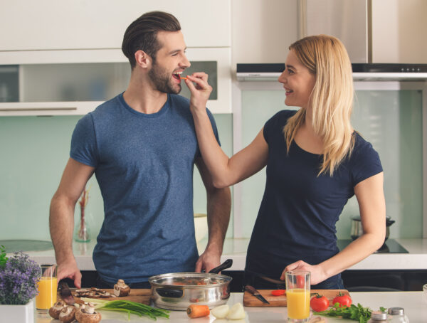 Symbolbild: Mann und Frau stehen in moderner Küche, Frau füttert Mann mit Karotte, auf Arbeitsfläche Gemüse, Kräuter und zwei Gläser Orangensaft.