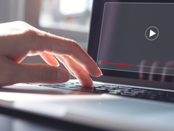 Symbolbild mit einer Hand, die auf die Tastatur eines Laptops klickt, auf dem Bildschirm ist das Play-Zeichen eines Videos