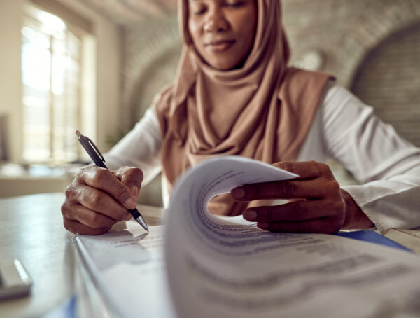 Symbolbild eine Fraue mit einem Kopftuch schreibt mit einem Stift auf Papierblättern
