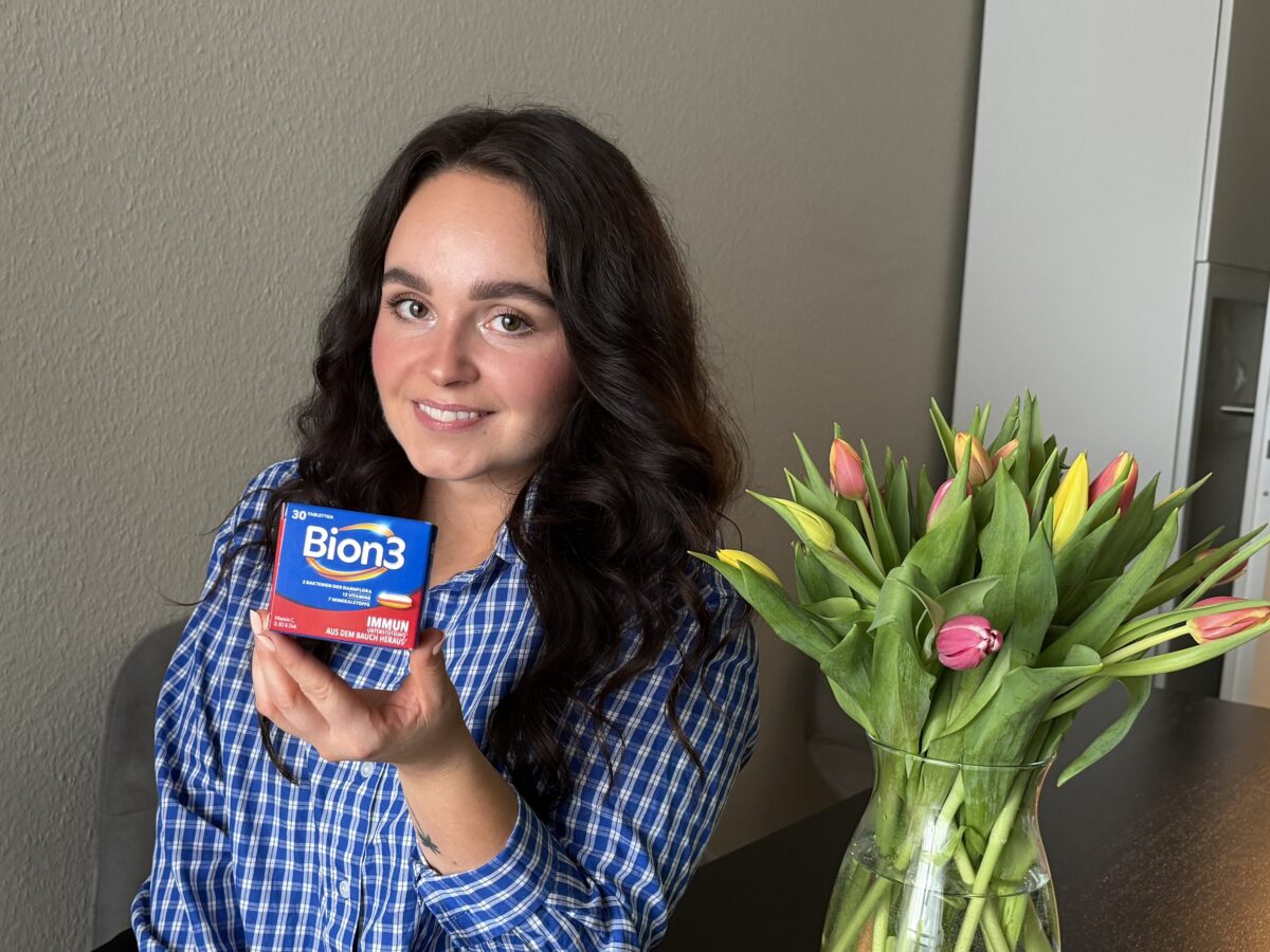Person hält eine Packung Bion3 in der Hand; auf dem Tisch steht ein Strauß bunter Tulpen in einem Innenraum.