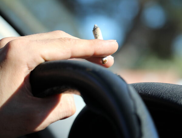 Symbolbild mit einer Hand am Steuer eines Autos mit einer Zigarette oder einem Joint in der Hand.
