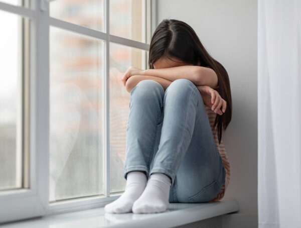 Symbolbild mit einem Mädchen, dass auf der Fensterbank sitzt und ihren Kopf auf ihren Beinen angelehnt hat mit verschränkten Armen.